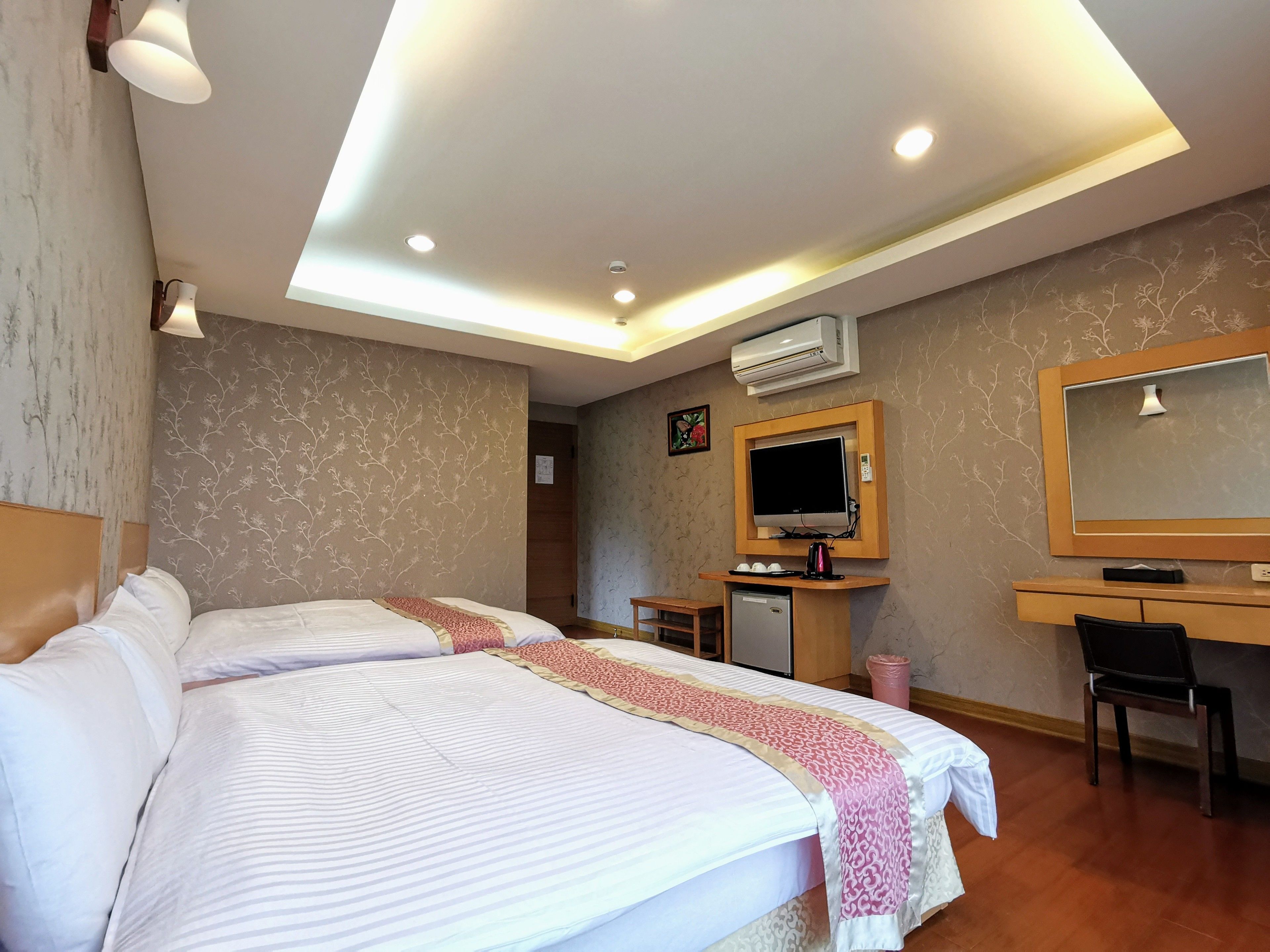 A hotel room with two beds, a desk, a chair, a television, and an air conditioning unit.