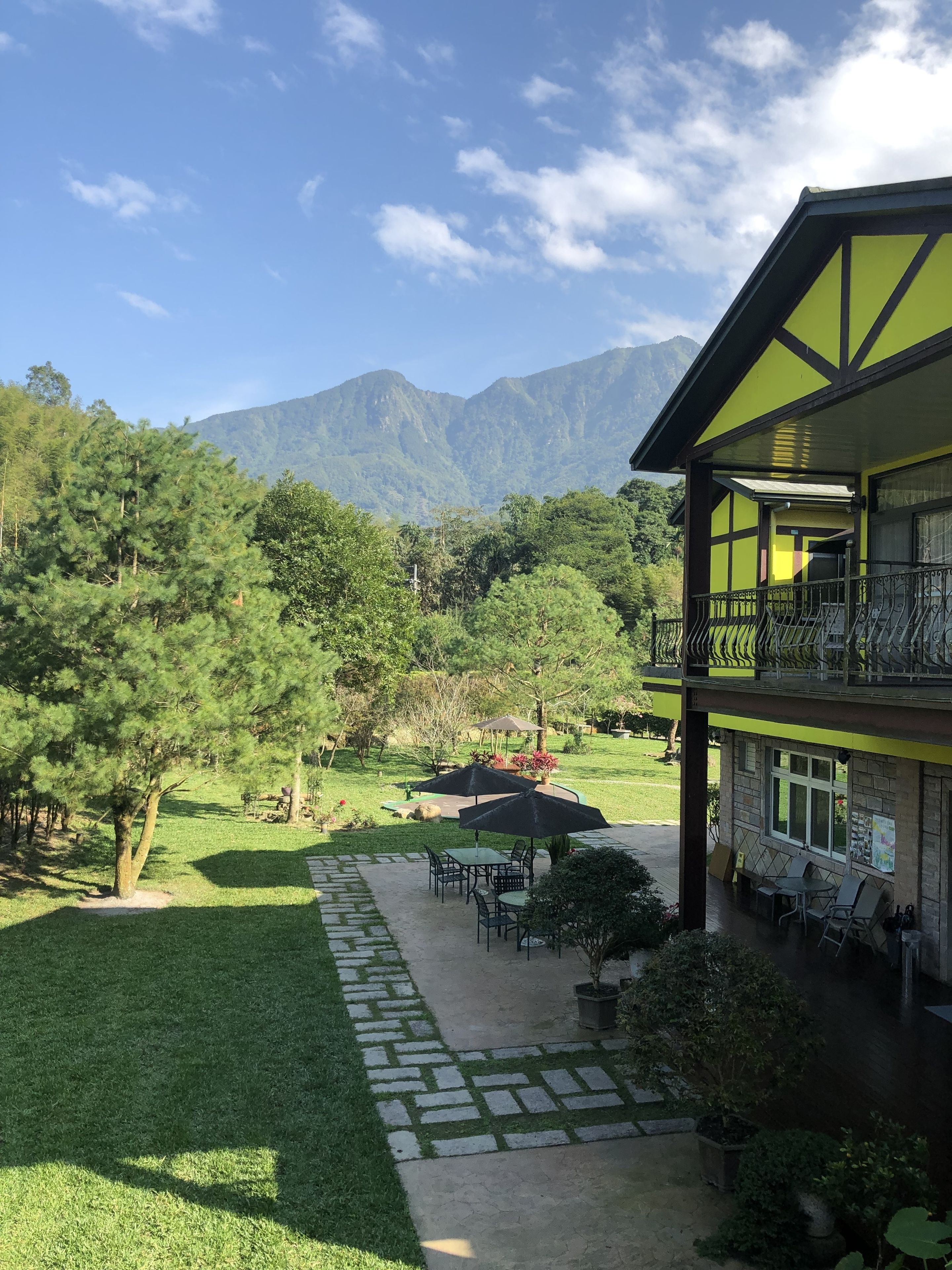 Ein Gebäude mit Balkon, das auf einen Garten mit Tischen und Stühlen blickt, umgeben von Bäumen und Bergen im Hintergrund.