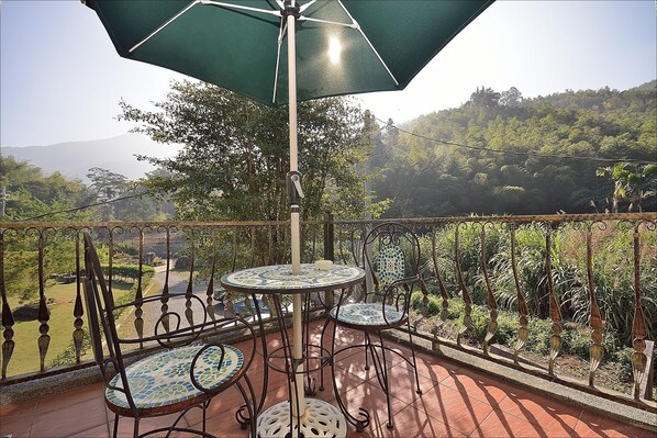 Un balcon avec une table et des chaises, un parasol vert, et une vue sur un paysage verdoyant avec des collines en arrière-plan.