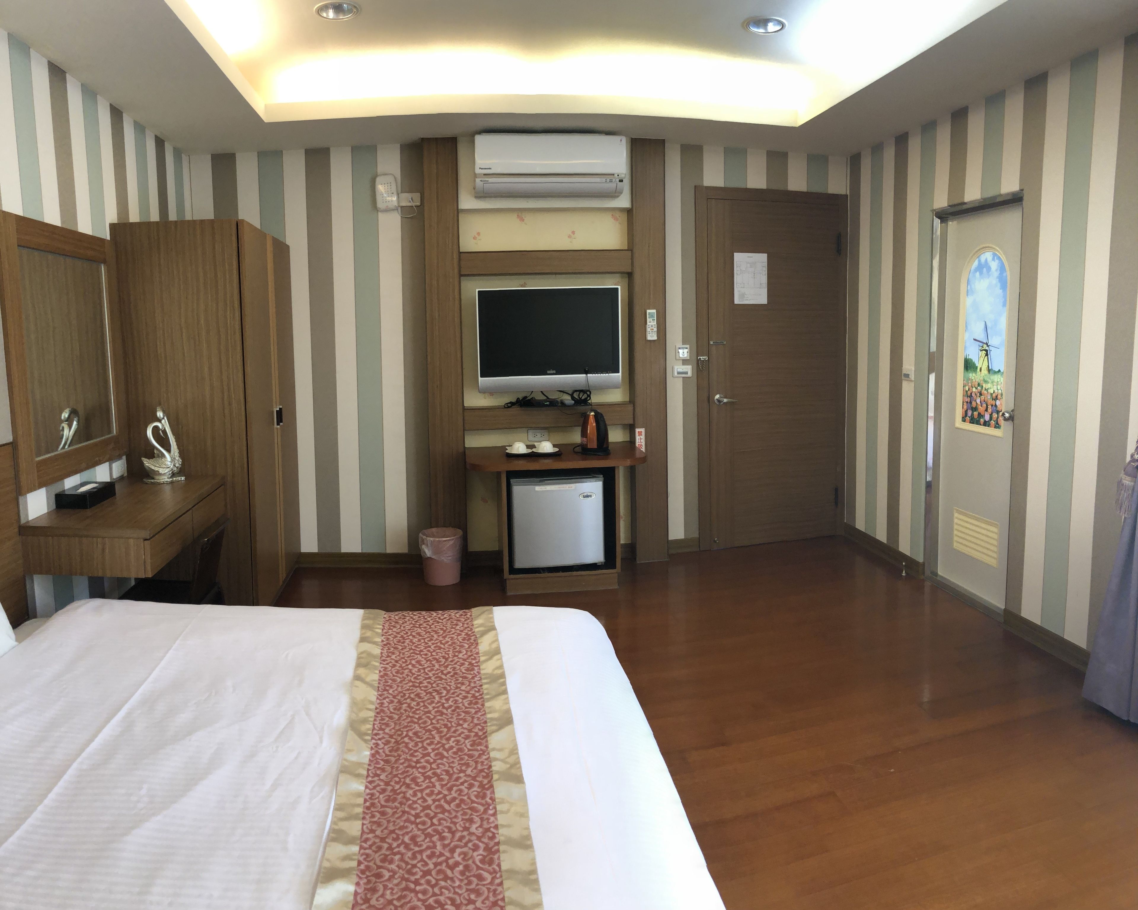 Habitación de hotel con cama, escritorio con computadora, televisor, refrigerador y puerta decorada con un cuadro.
