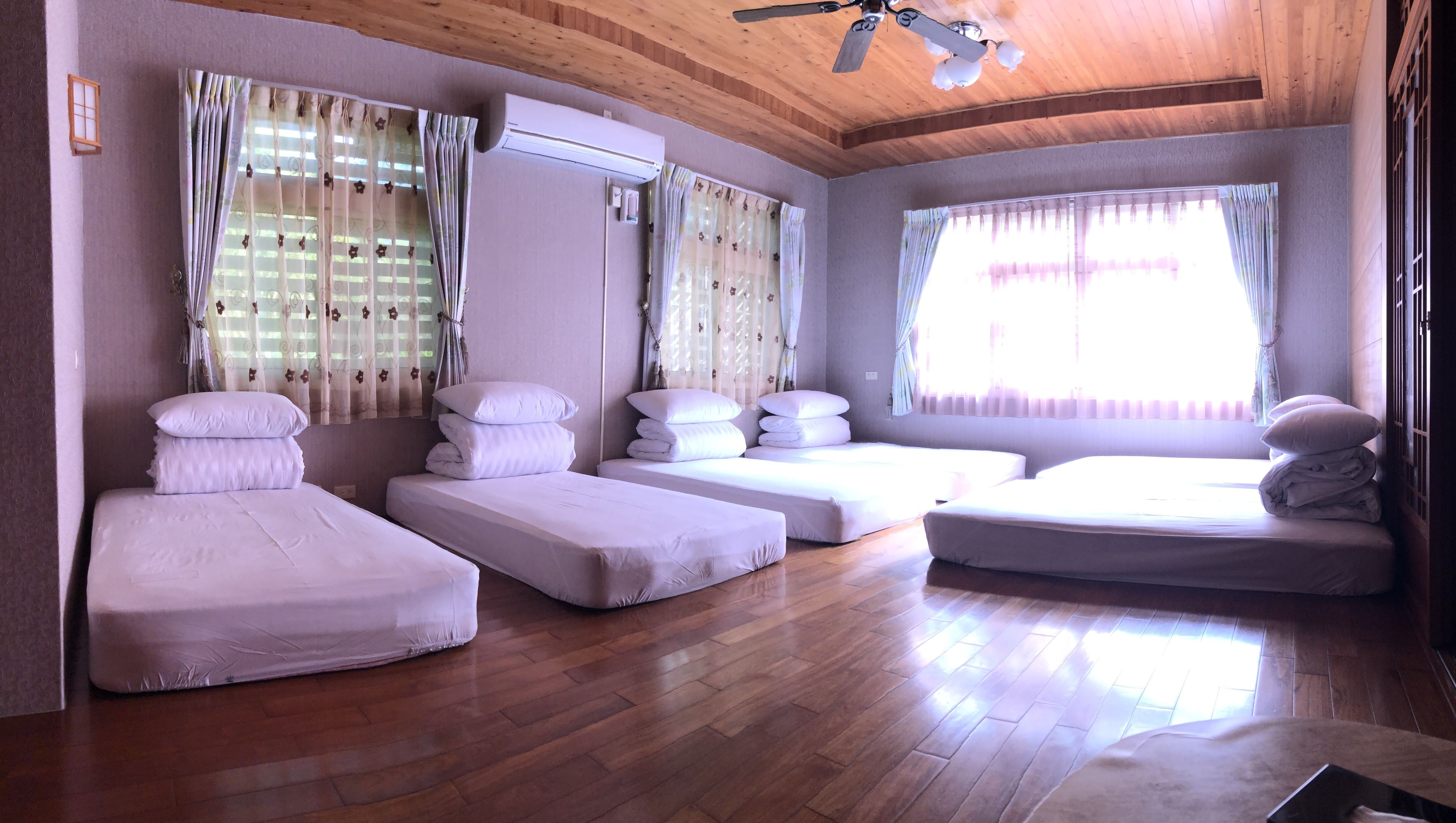 A room with four beds, wooden flooring, a ceiling fan, and large windows with curtains.