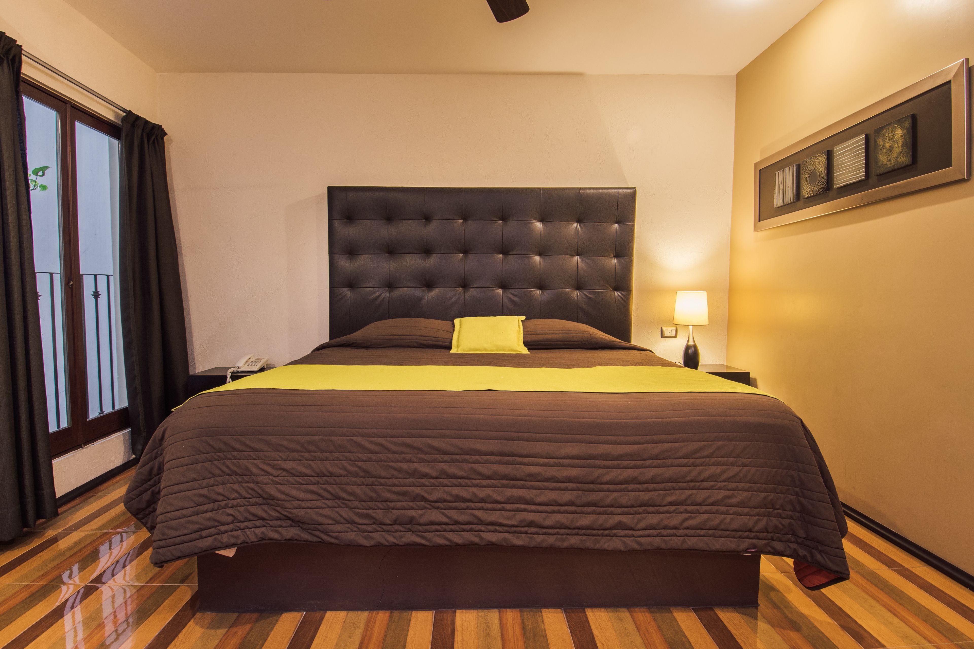Ein großes Bett mit einem gestopften Kopfteil, einem gelben Kissen und einer braunen Bettdecke. Ein Nachttisch mit einer Lampe und einem Telefon steht daneben. Der Raum hat Holzboden und ein Fenster mit Vorhängen.