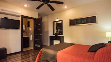 Quarto de hotel com cama, ventoinha no teto, TV, banheiro com espelho e obra de arte pendurada na parede.