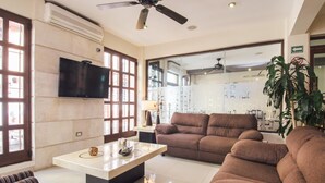 A spacious living area with a large flat-screen TV, comfortable sofas, a coffee table, and a glass partition separating the living space from a fitness area.