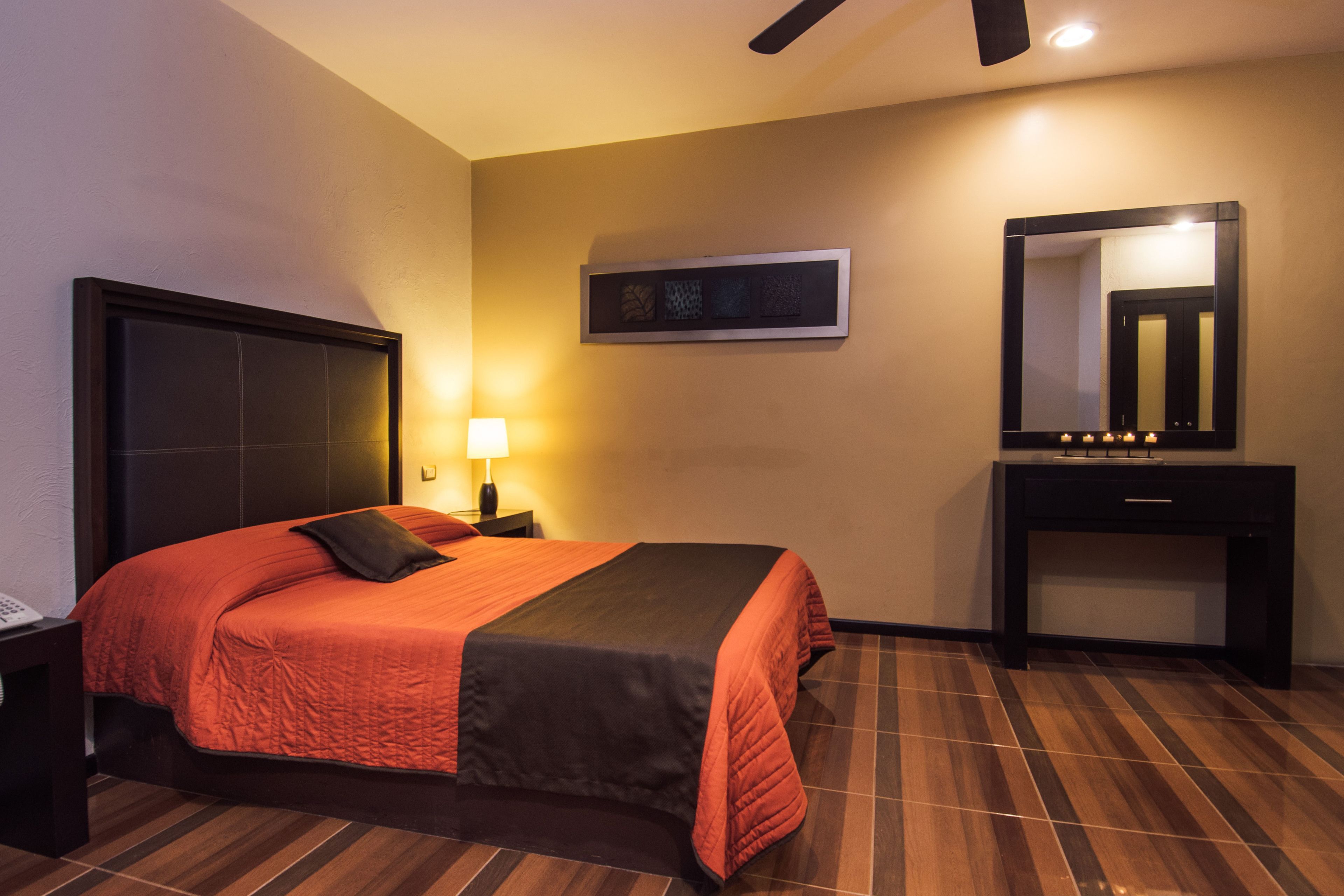 Habitación de hotel con cama, mesita de noche, lámpara, cómoda, espejo y una obra de arte enmarcada en la pared.