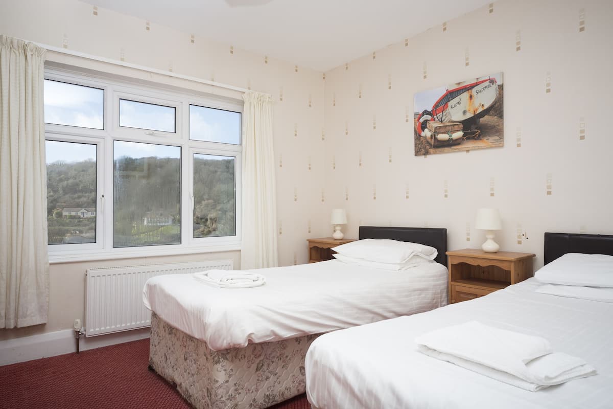A hotel room with two beds, a window with curtains, a radiator, a painting on the wall, and a small wooden nightstand with a lamp.