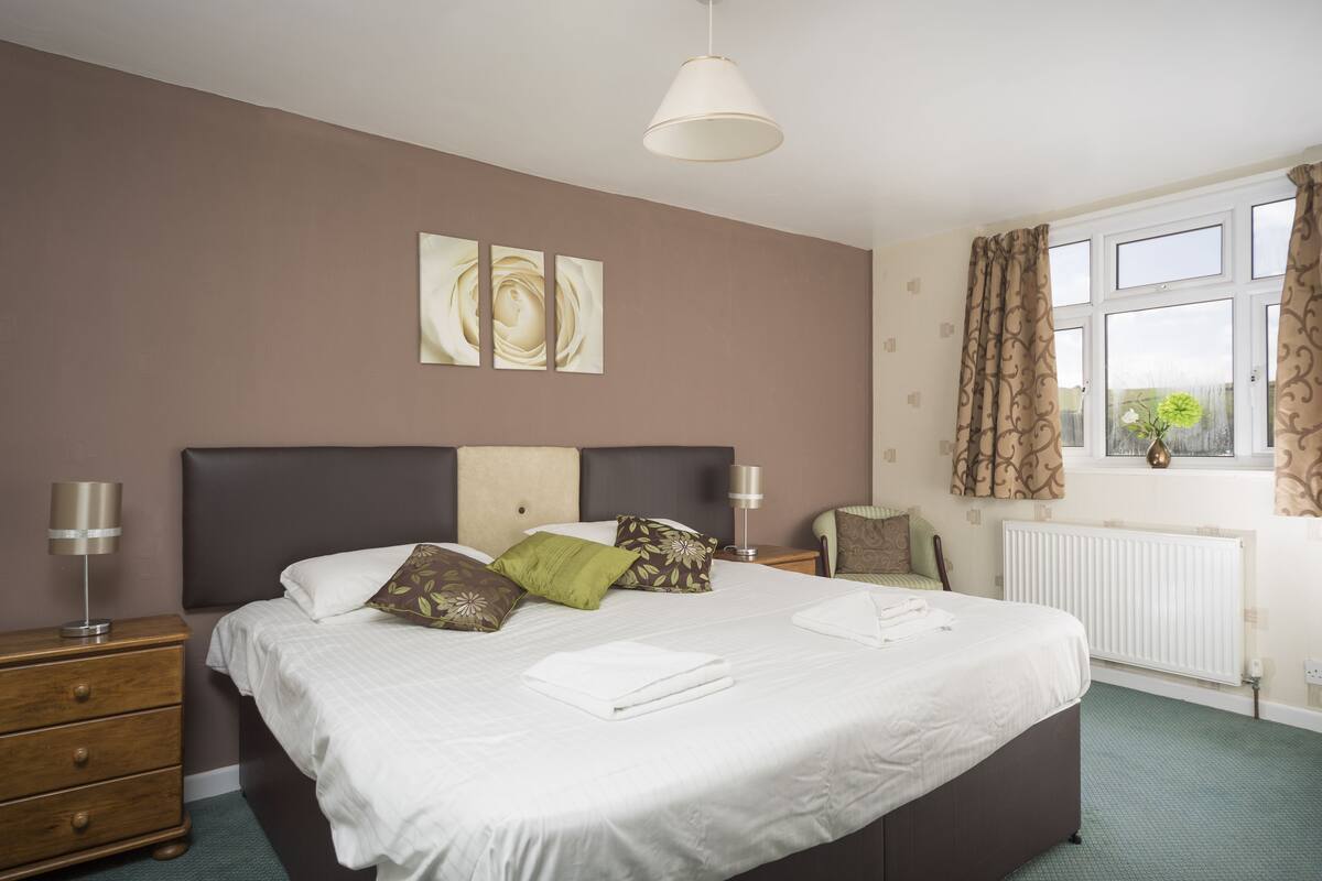 A neatly made bed with white linens, a brown headboard, and decorative pillows. There is a bedside table with a lamp, a window with curtains, and a radiator.