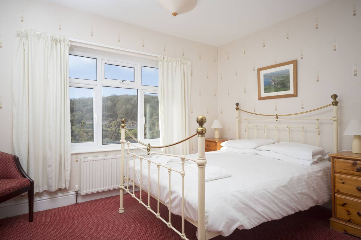 A neatly arranged bedroom with a large window, a bed with white linens, a wooden nightstand, and a red carpeted floor.