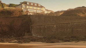 A coastal building with a flat roof and multiple windows, situated on a sandy beach with a stone wall in the foreground. - The Sun Bay Hotel & Restaurant (Kingsbridge)