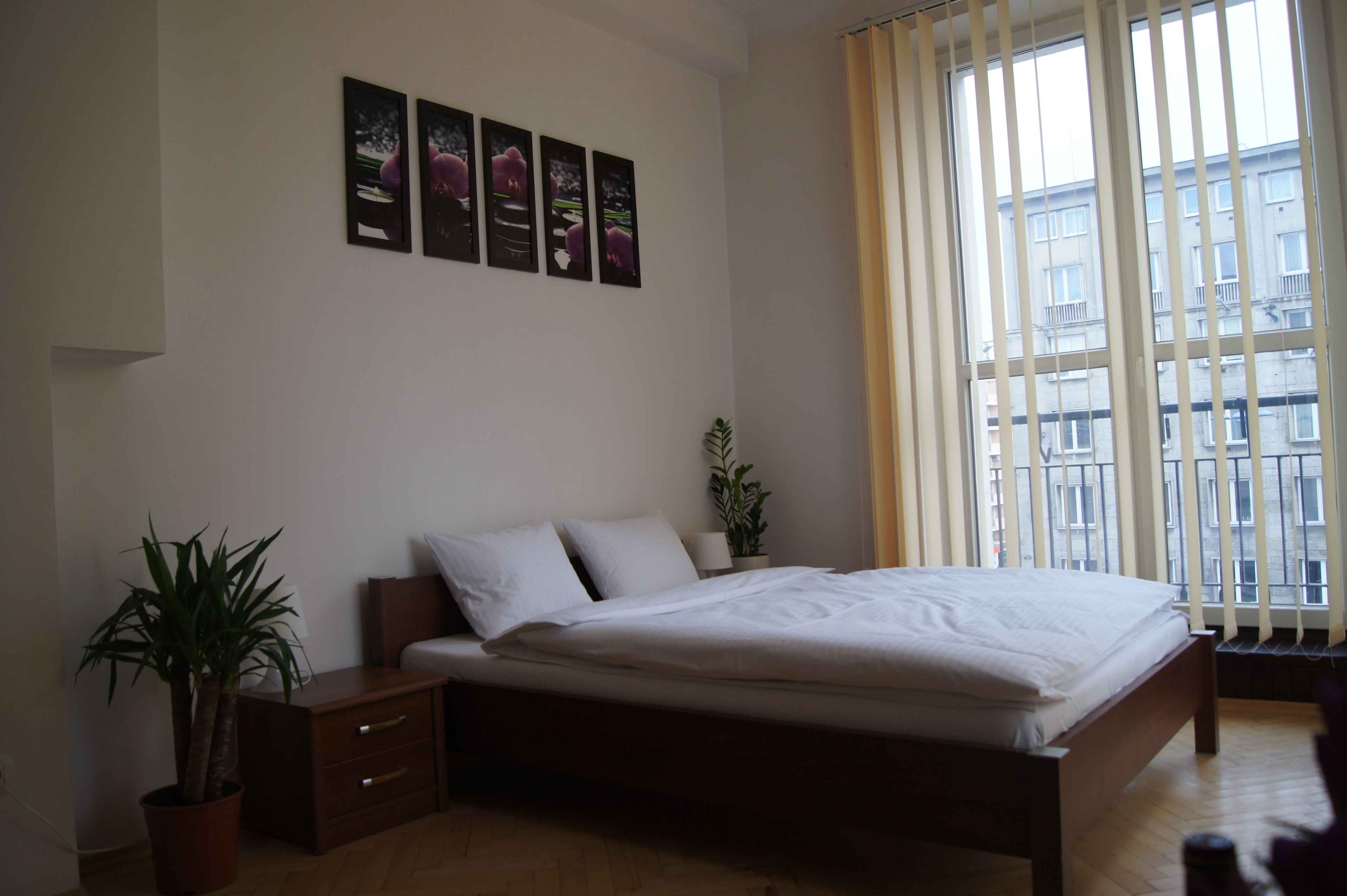 Quarto com cama, mesa de cabeceira, planta em vaso e janela com cortinas.