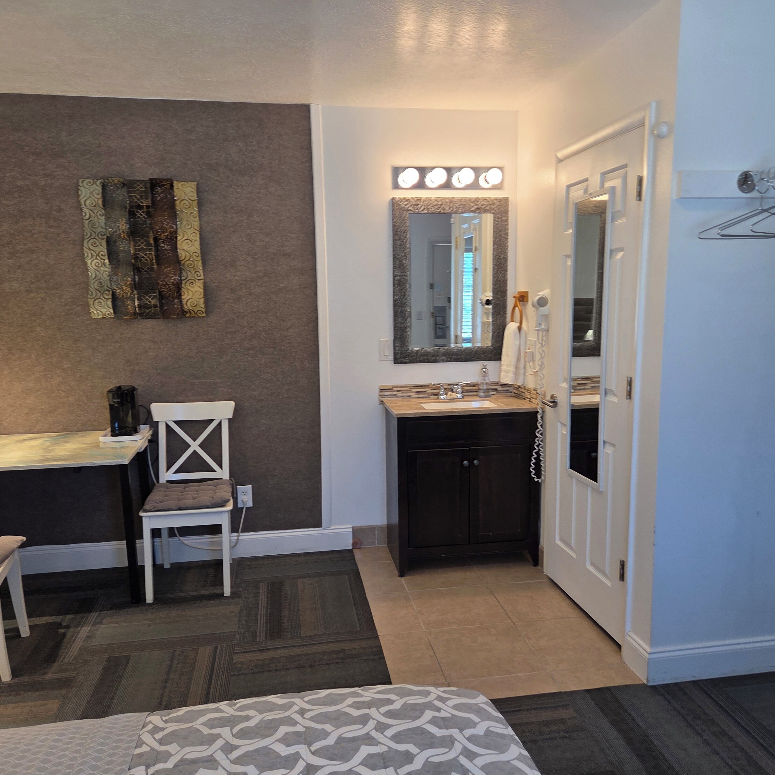 A bedroom with a bed, a desk, a chair, a vanity with a mirror, and a door.