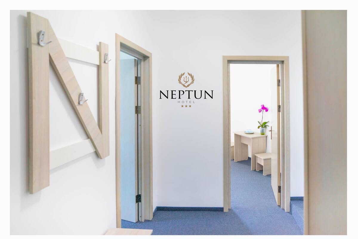 Un pasillo con una decoración mural en forma de N de madera, un letrero del Hotel Neptuno y una vista parcial de una habitación con escritorio y silla.