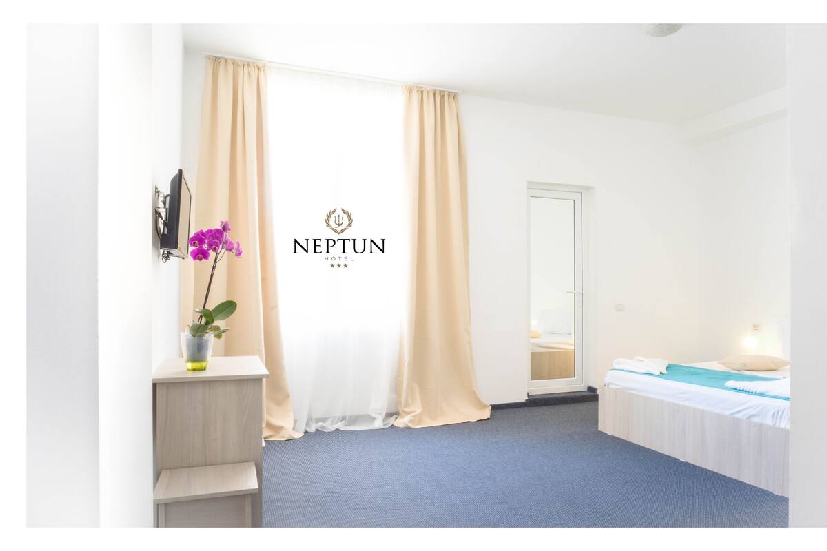 Habitación de hotel con cama, una mesita con una toalla, mesita de noche, espejo y ventana con cortinas.