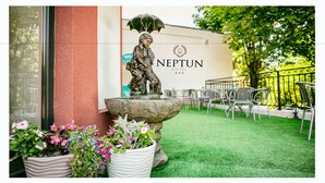 An outdoor seating area with a statue of a child under an umbrella, potted plants, and a sign for 'NEPTUN HOTEL'.