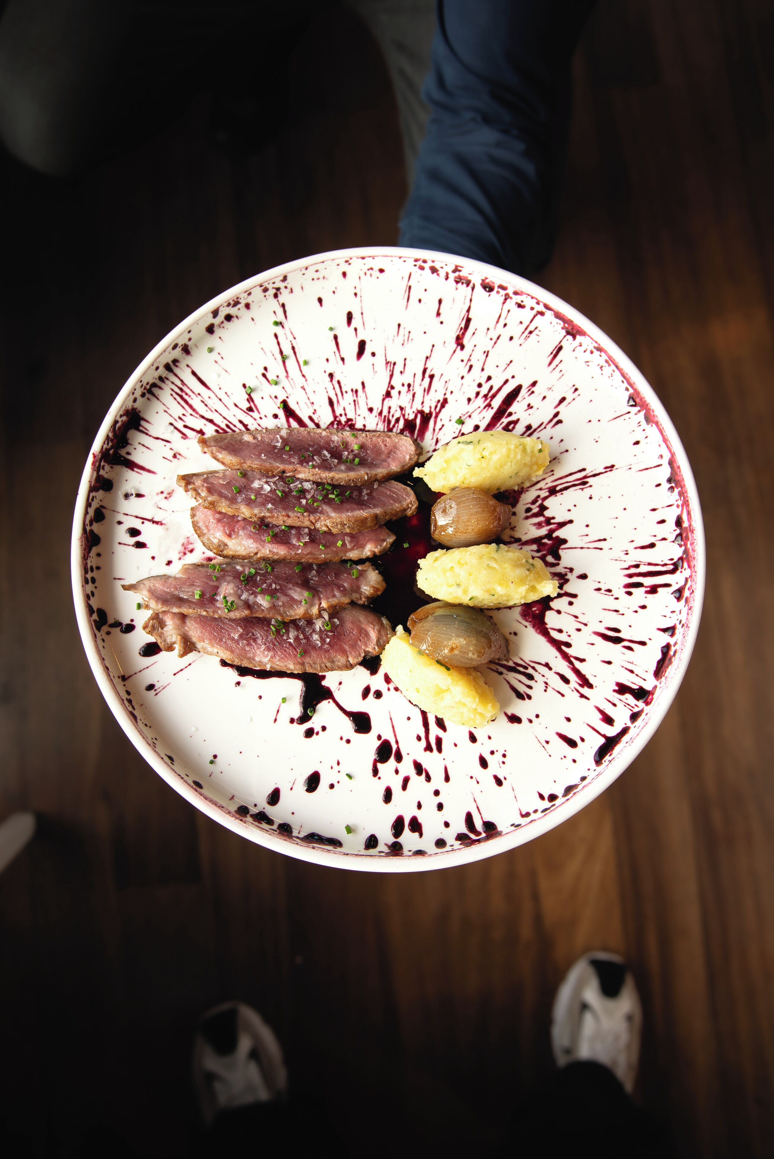 A plate of food with meat and potatoes, held by a person standing on a wooden floor.