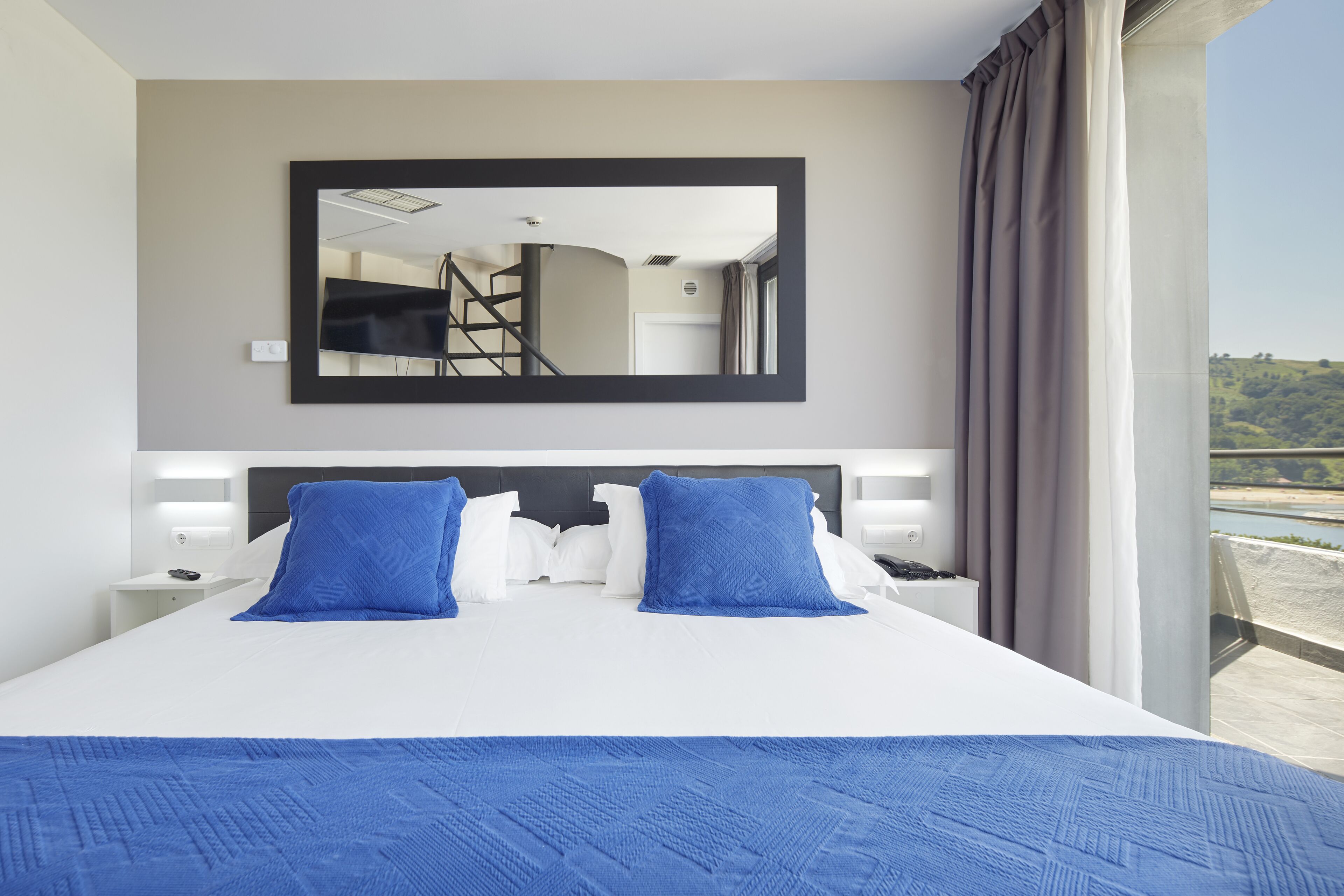 A hotel room with a large bed, a mirror, a nightstand, and a view of a balcony.