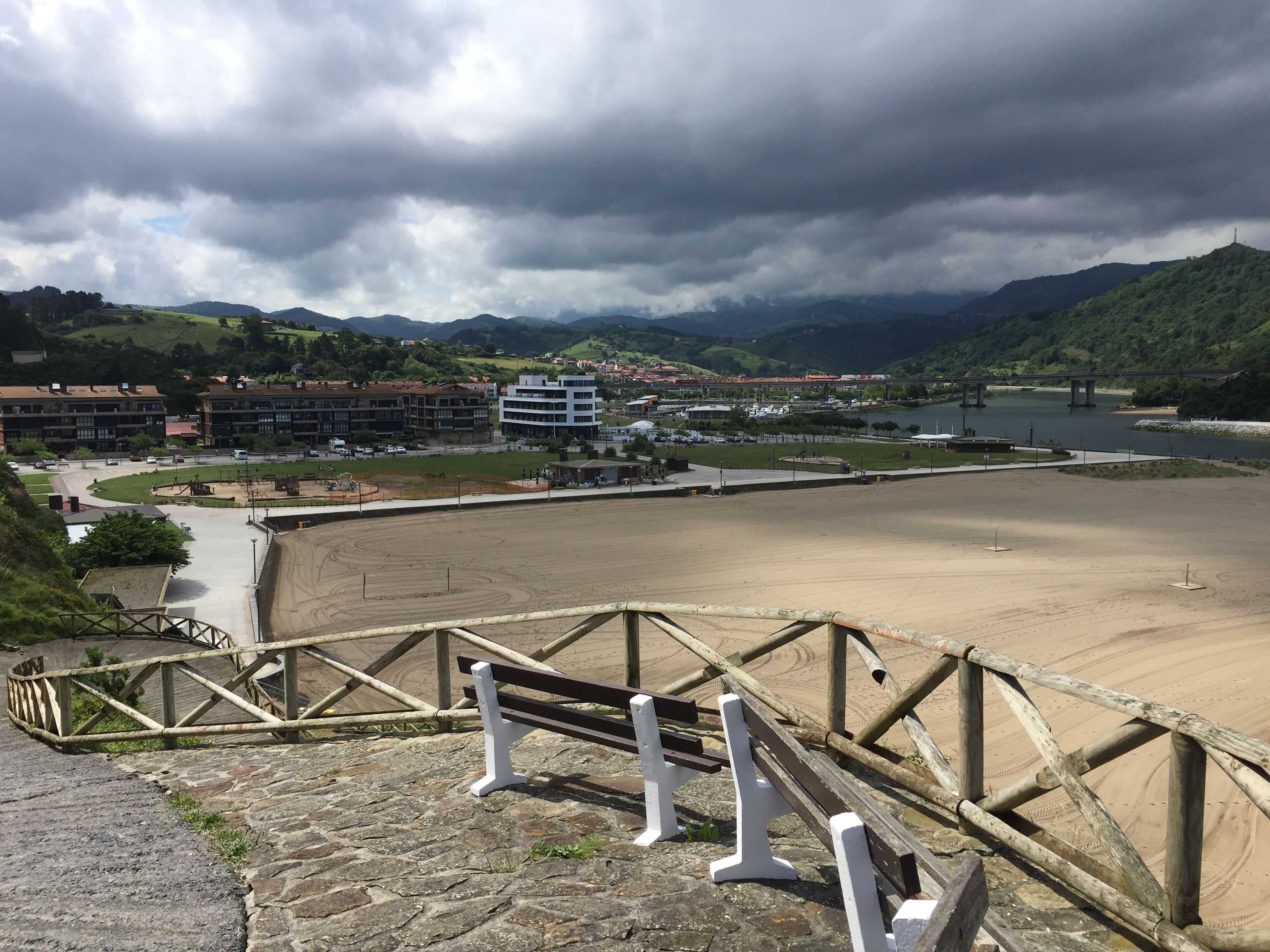 A coastal view with a sandy beach, a wooden staircase, and a view of a town and mountains in the background.