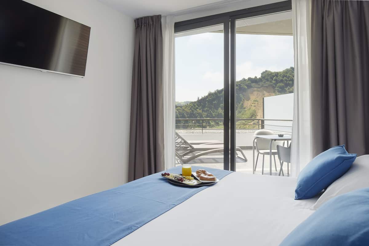 Habitación de hotel con cama, bandeja de desayuno, televisión y balcón con vistas a las colinas.