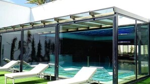 Zona de piscina con paredes de vidrio, dos sillones de descanso y una palmera al fondo.