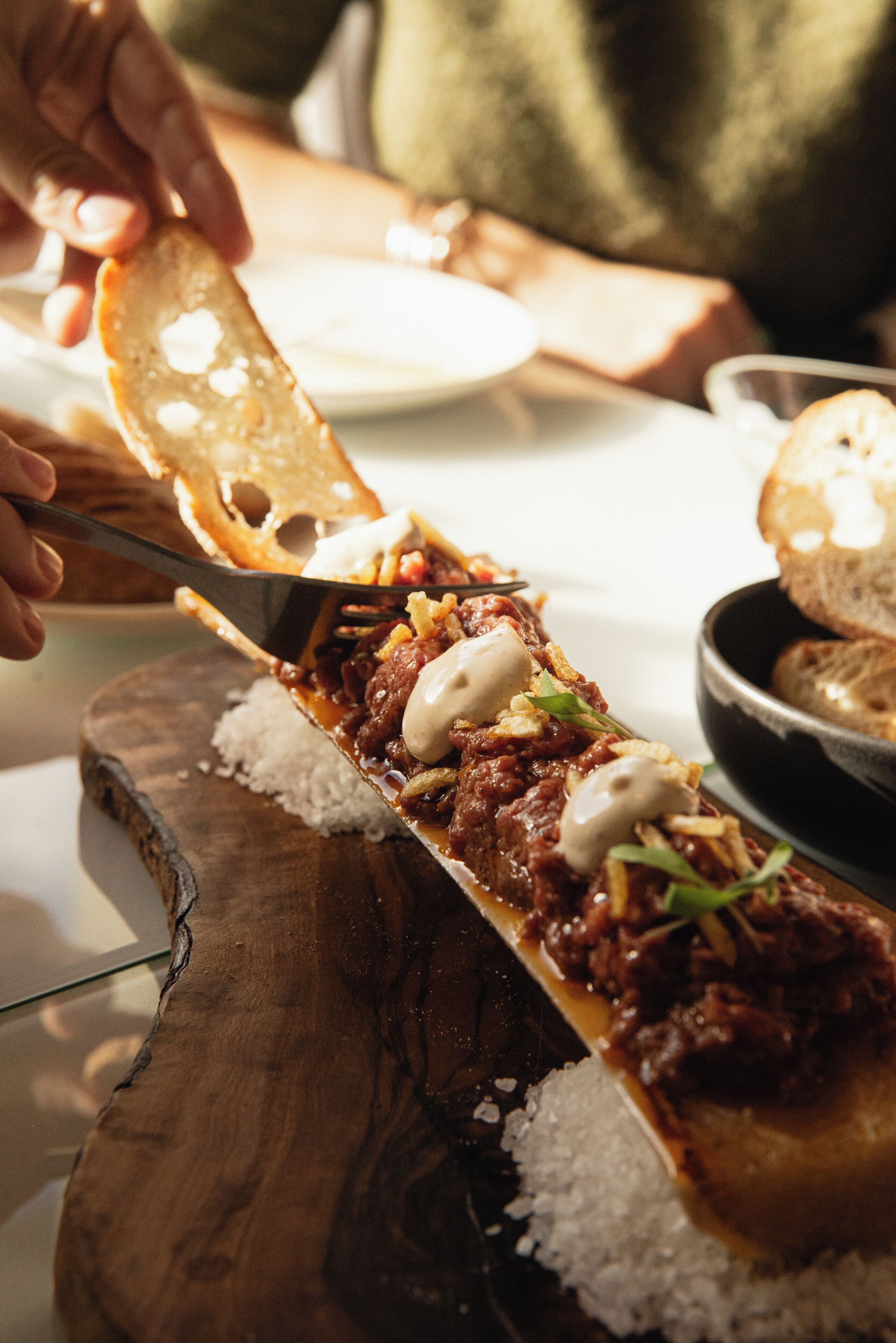 A wooden serving board with a dish of meat and vegetables, a bowl of bread, and a person cutting the bread.