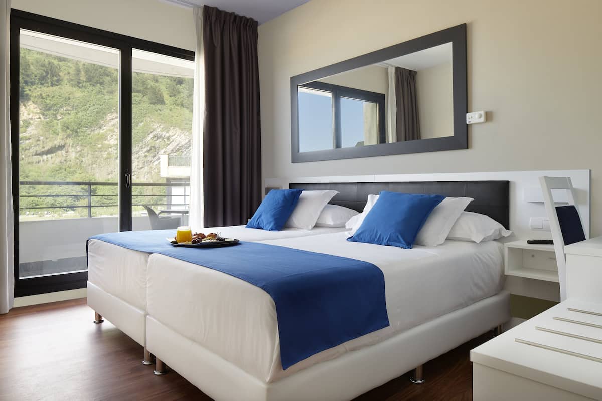 Habitación de hotel con una cama grande, una mesita de noche, un espejo y vistas a un paisaje montañoso por la ventana.