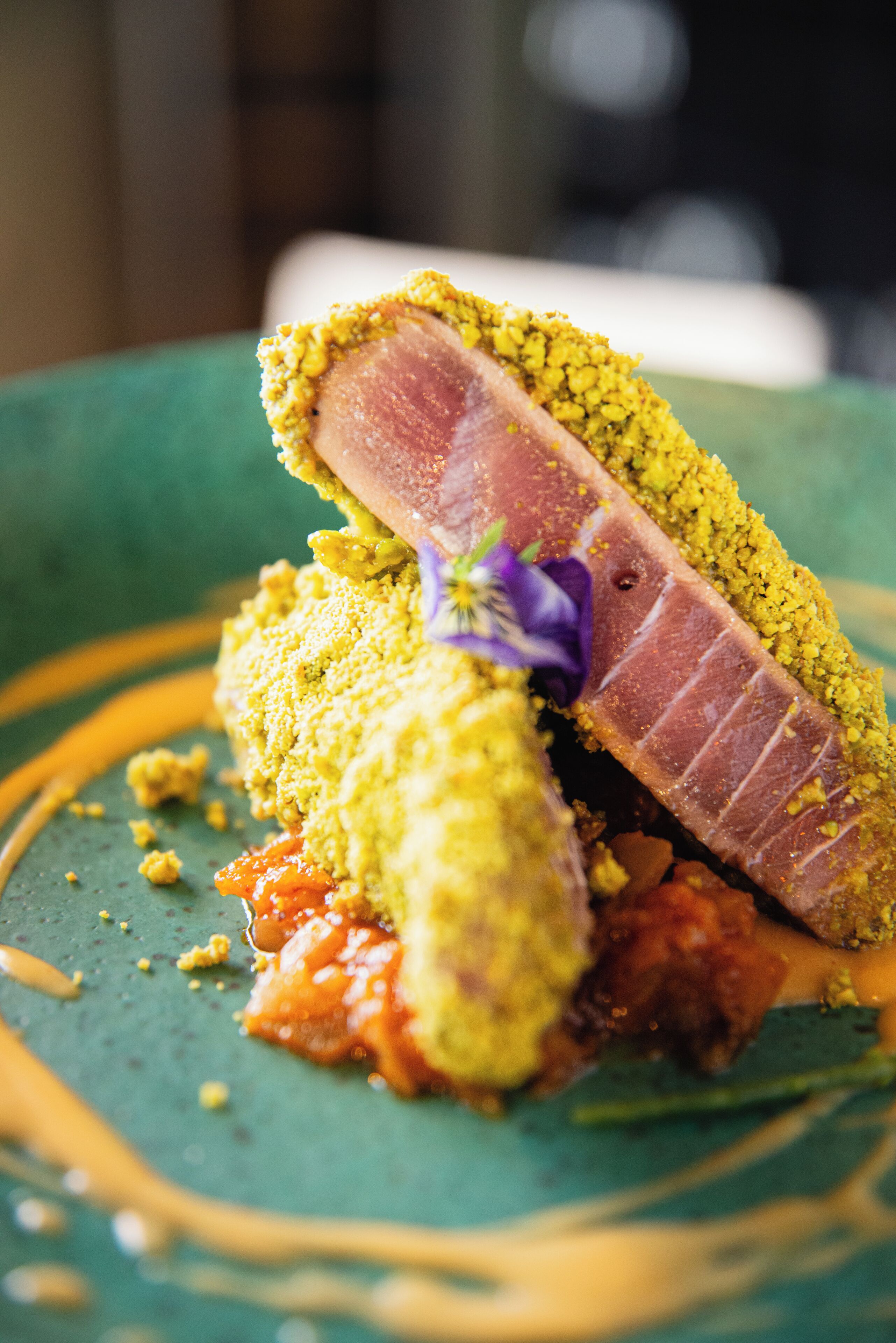 A close-up of a plated dish with a slice of meat, crumbly topping, and a decorative flower.