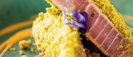 A close-up of a plated dish with a slice of meat, crumbly topping, and a decorative flower.