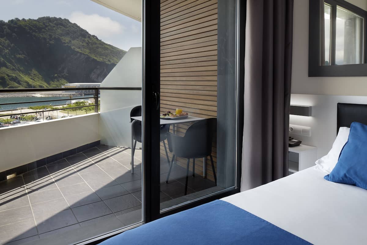 Una habitación de hotel moderna con balcón, una cama, una mesita con una silla y vistas a un paisaje montañoso.