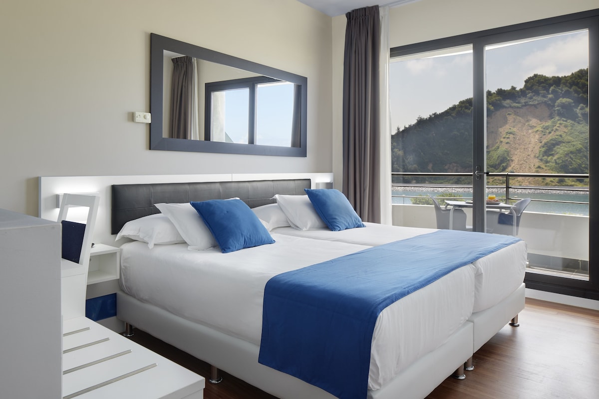 Una habitación de hotel moderna con una cama grande, una mesita de noche, un espejo y vistas a un balcón con mobiliario exterior.