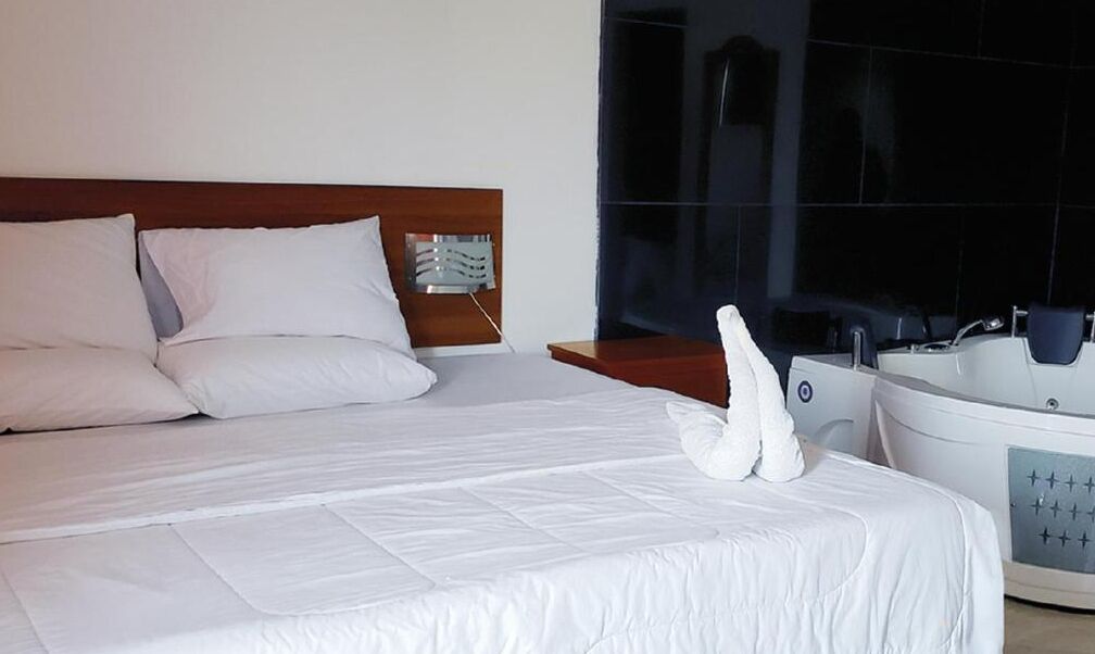A hotel room with a large bed, a wooden headboard, a bedside table, a modern bathroom with a bathtub, and a large mirror.