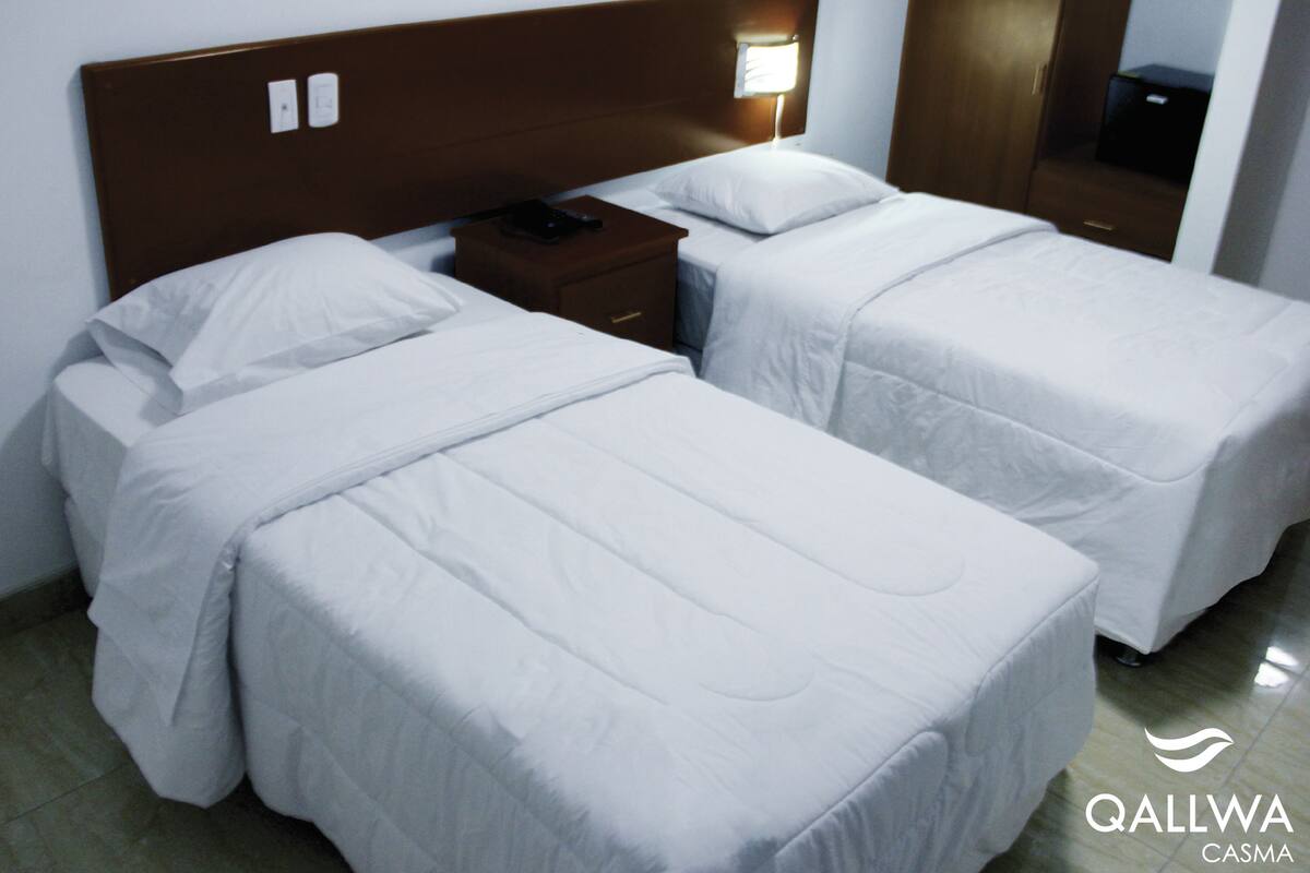 A hotel room with two single beds, a wooden headboard, a bedside table with a lamp, and a mirror.