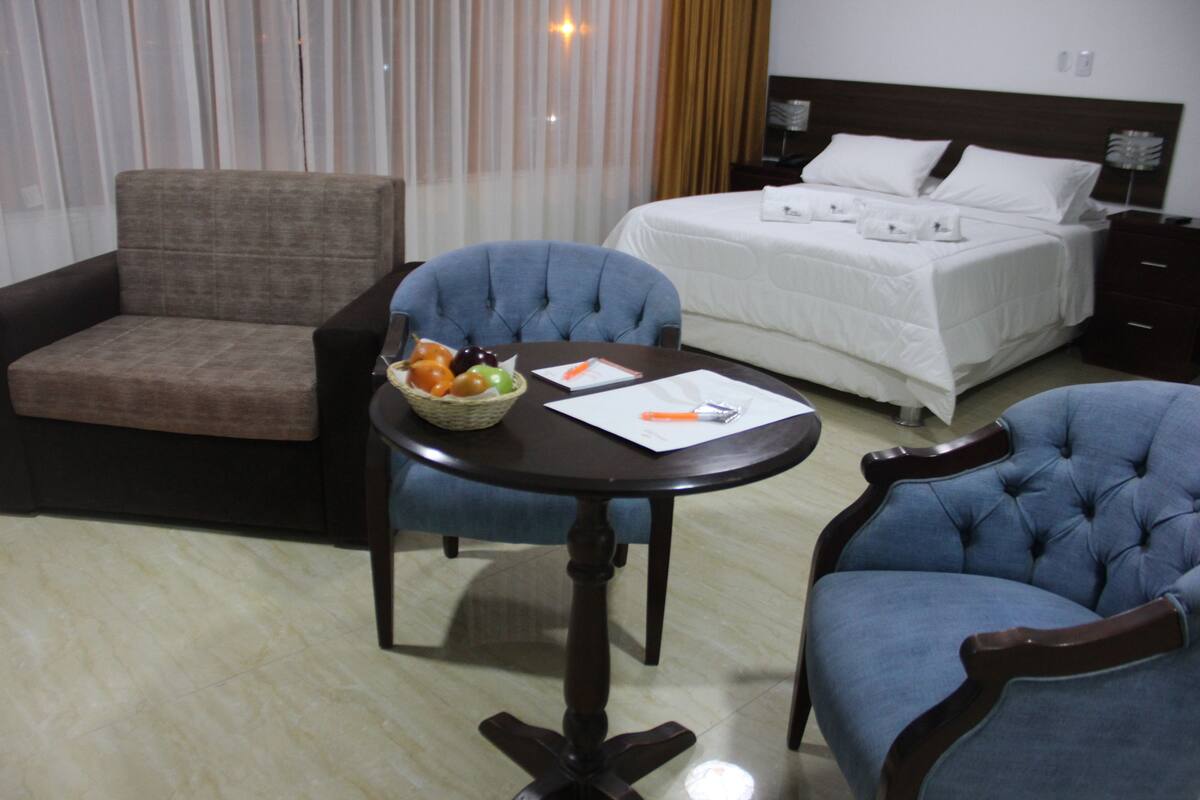 A hotel room with a bed, a dining table with chairs, a small sofa, and a fruit basket.