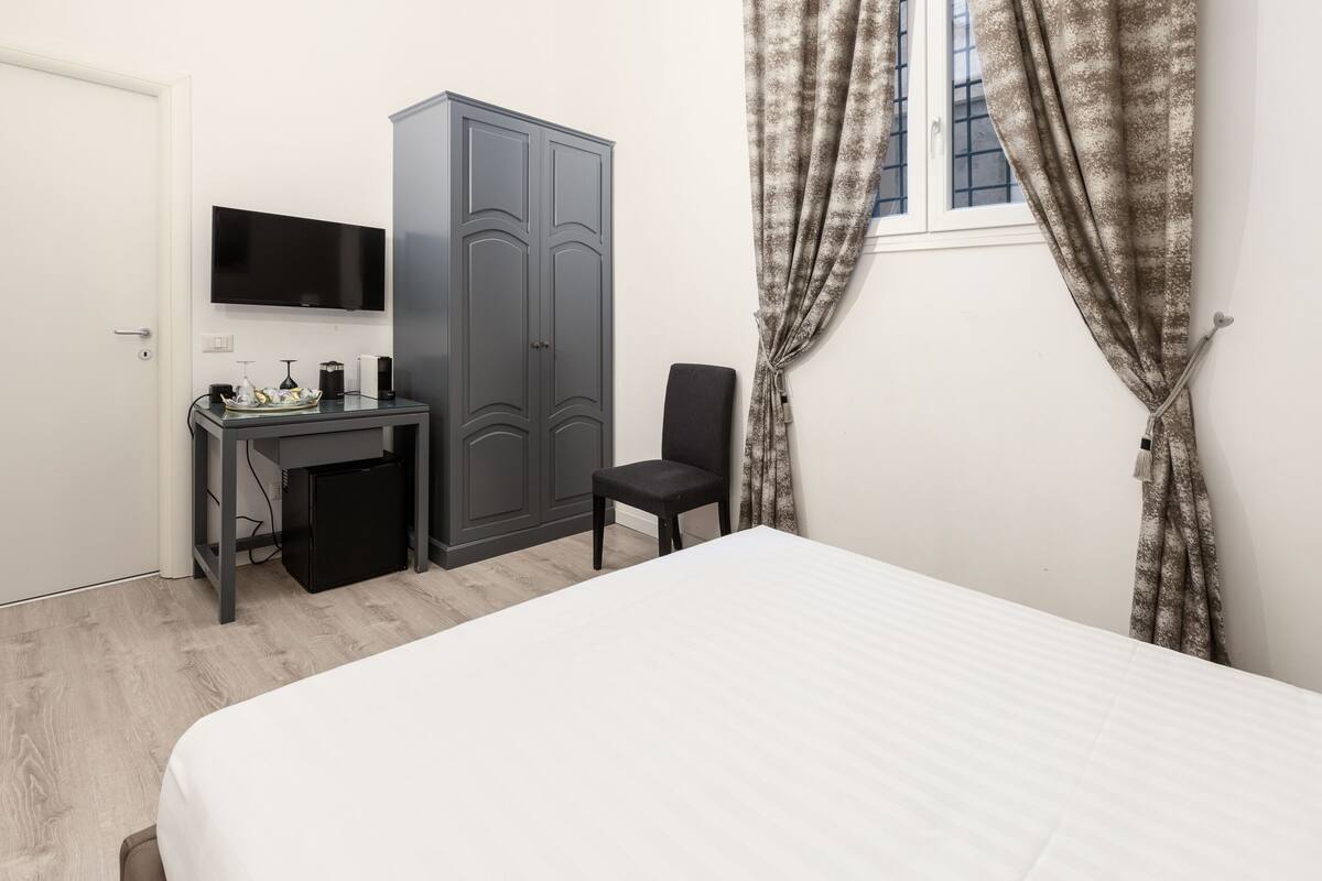 A bedroom with a bed, a desk with a chair, a wardrobe, a television, and a window with curtains.