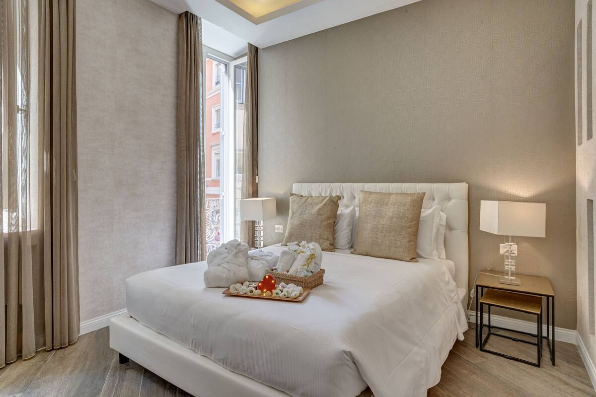 A hotel room with a large bed, a bedside table with a lamp, and a tray with towels and a heart-shaped ornament.