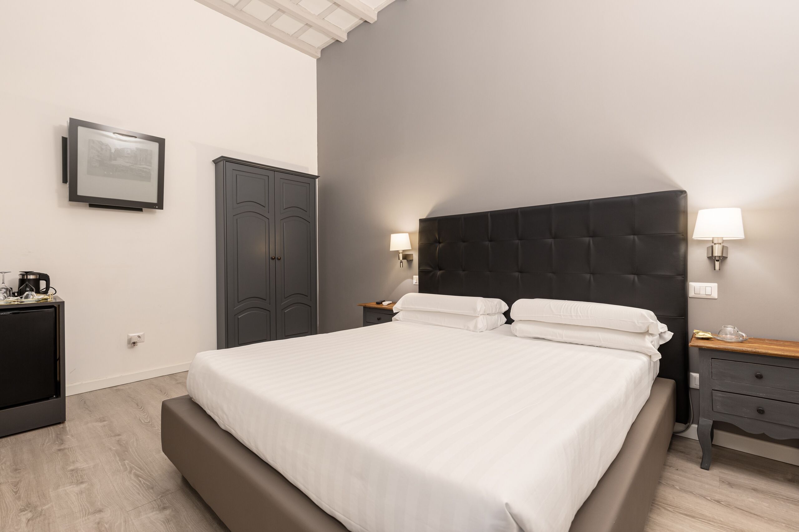 A modern hotel room with a large bed, a wardrobe, a bedside table with a lamp, and a framed picture on the wall.