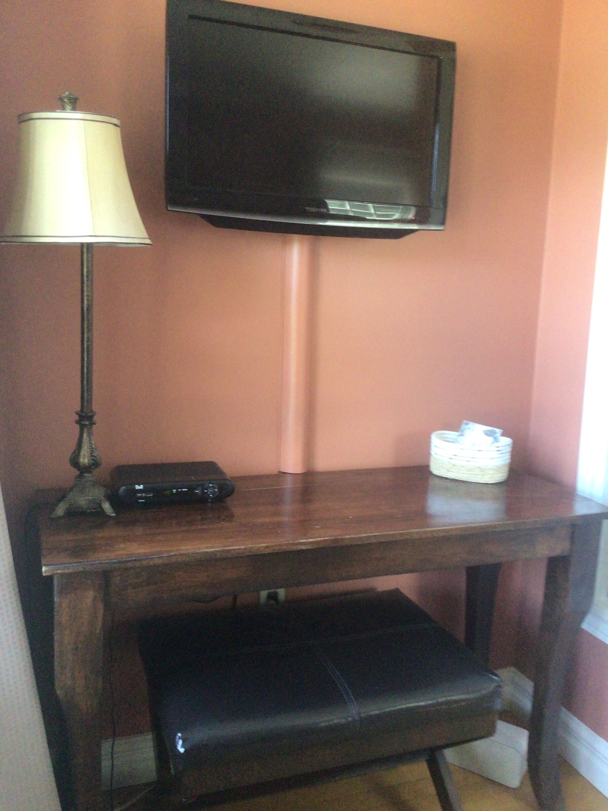 A room with a television mounted on the wall, a desk with a lamp, a remote control, and a small basket.
