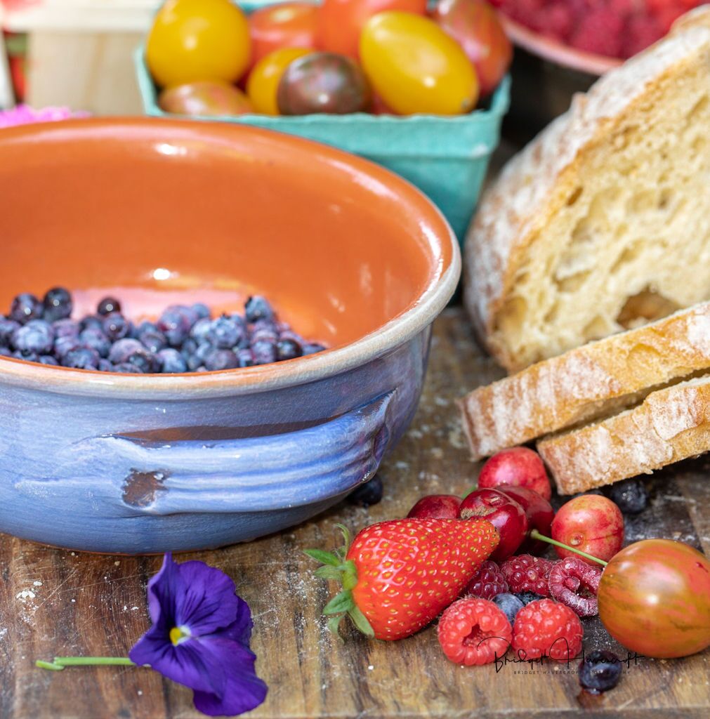 A rustic wooden table with a blue ceramic bowl filled with blueberries, a strawberry, raspberries, and a plum. There is also a slice of bread and a purple flower on the table.