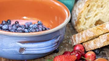 A rustic wooden table with a blue ceramic bowl filled with blueberries, a strawberry, raspberries, and a plum. There is also a slice of bread and a purple flower on the table.
