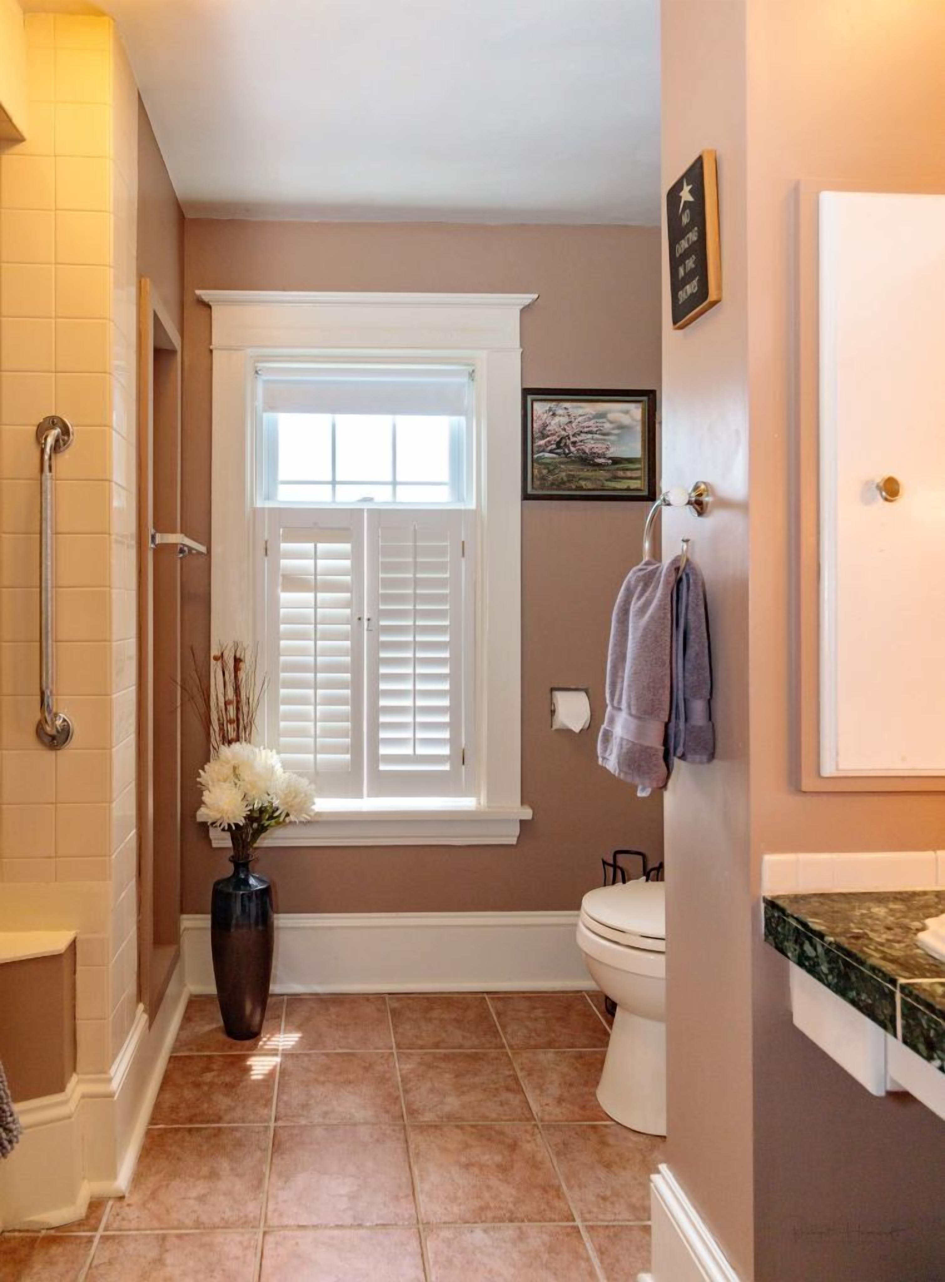 A bathroom with a toilet, a sink, a window with shutters, a vase with flowers, and a towel rack.