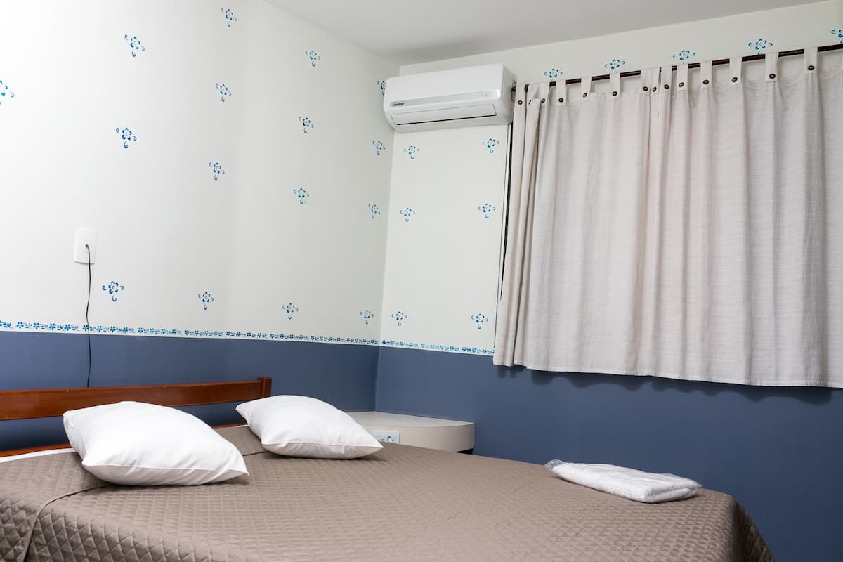 A bedroom with a bed, two pillows, a bedside table, and an air conditioning unit.