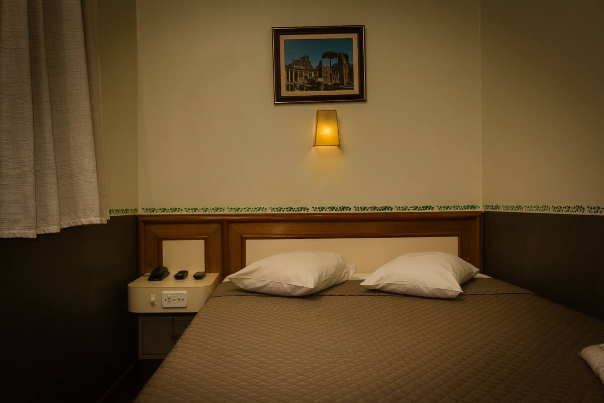 A hotel room with a bed, two pillows, a nightstand with a lamp, a remote control, and a wall-mounted picture.