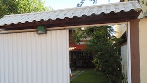 A covered outdoor area with a corrugated metal wall, a tiled roof, and a grassy lawn. - Hostel Praia Brava (Arraial do Cabo)