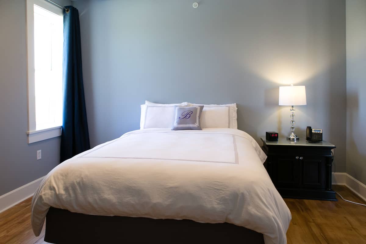 Un lit bien fait, avec du linge de lit blanc, une table de chevet avec une lampe et une petite table de nuit avec une horloge.
