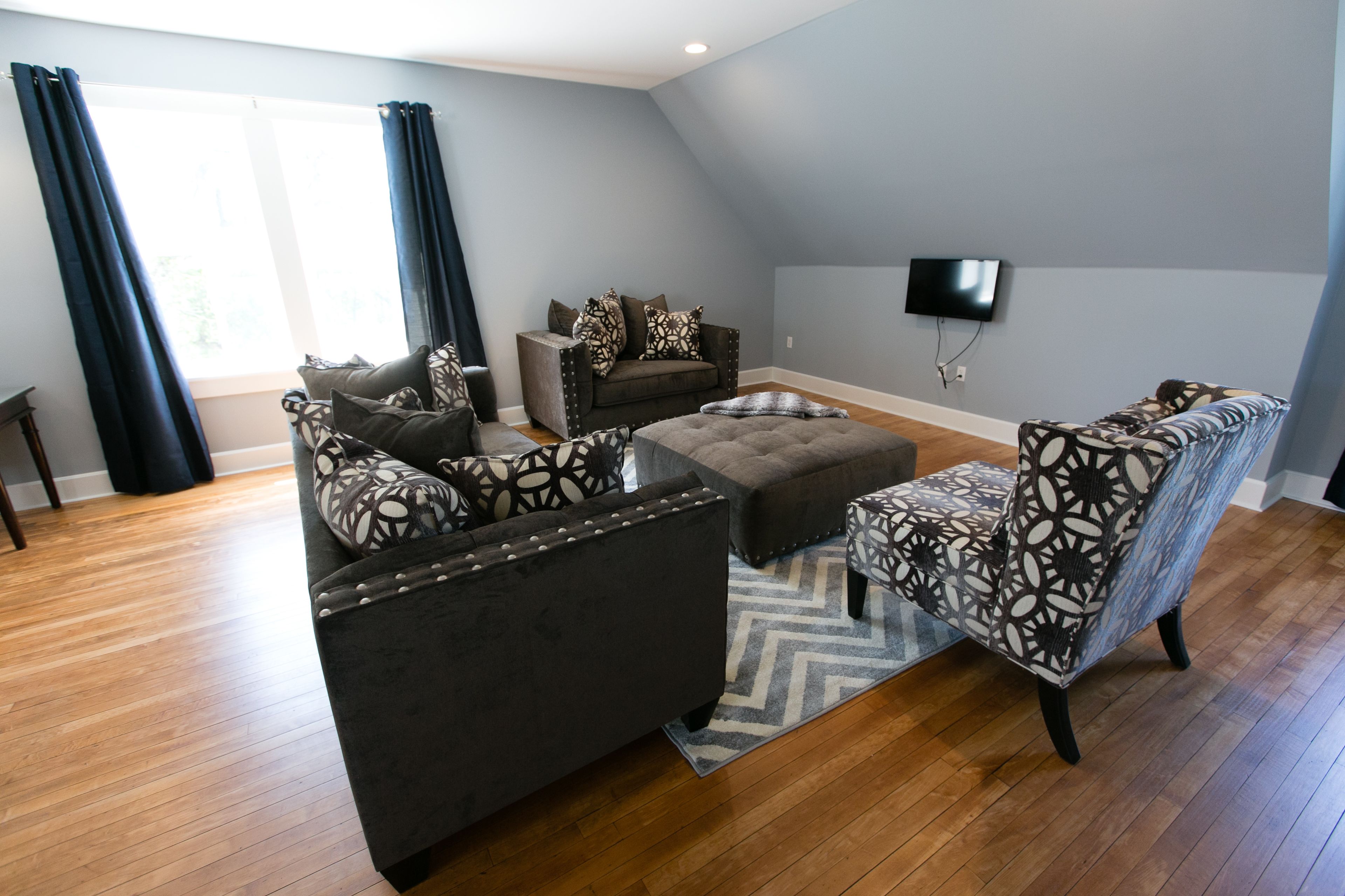 A living room with a sofa, armchair, ottoman, and a television mounted on the wall.