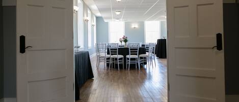 An open doorway leading to a room with a wooden floor, white doors, and a table with chairs arranged for a meeting or event.