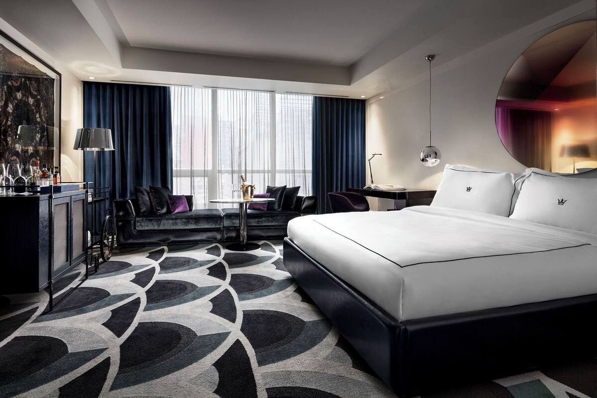 A modern hotel room with a large bed, a desk, a sofa, and a minibar.