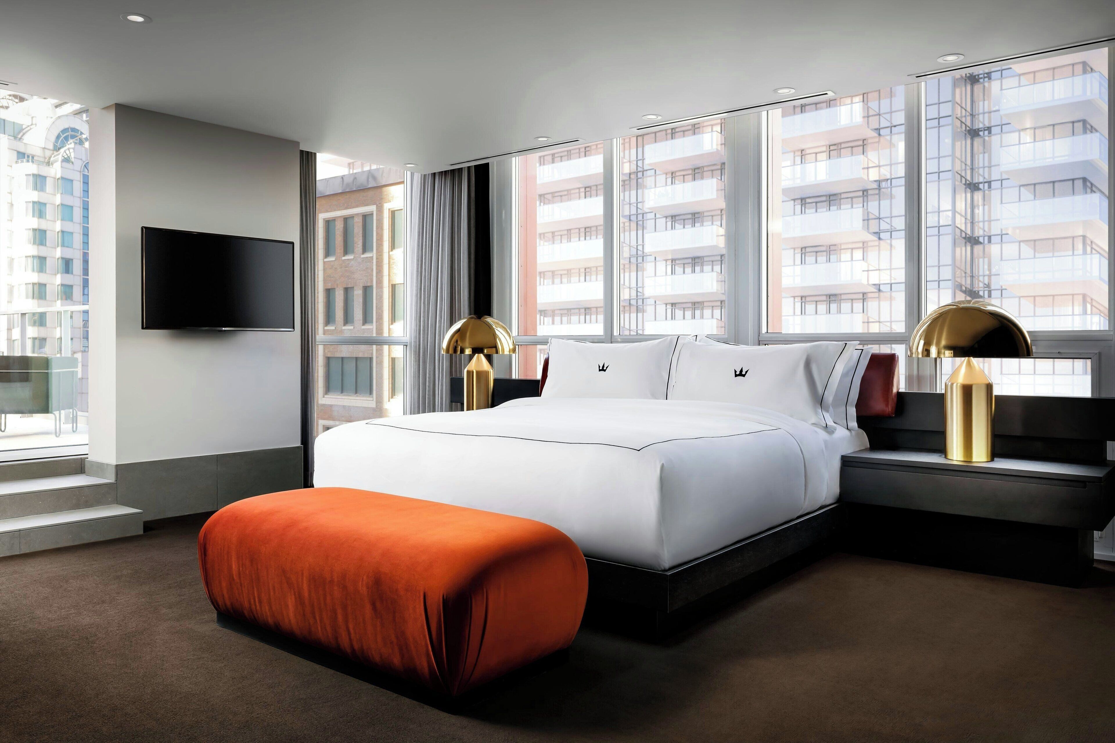 A modern hotel room with a large bed, a flat-screen TV, and a view of the cityscape through large windows.