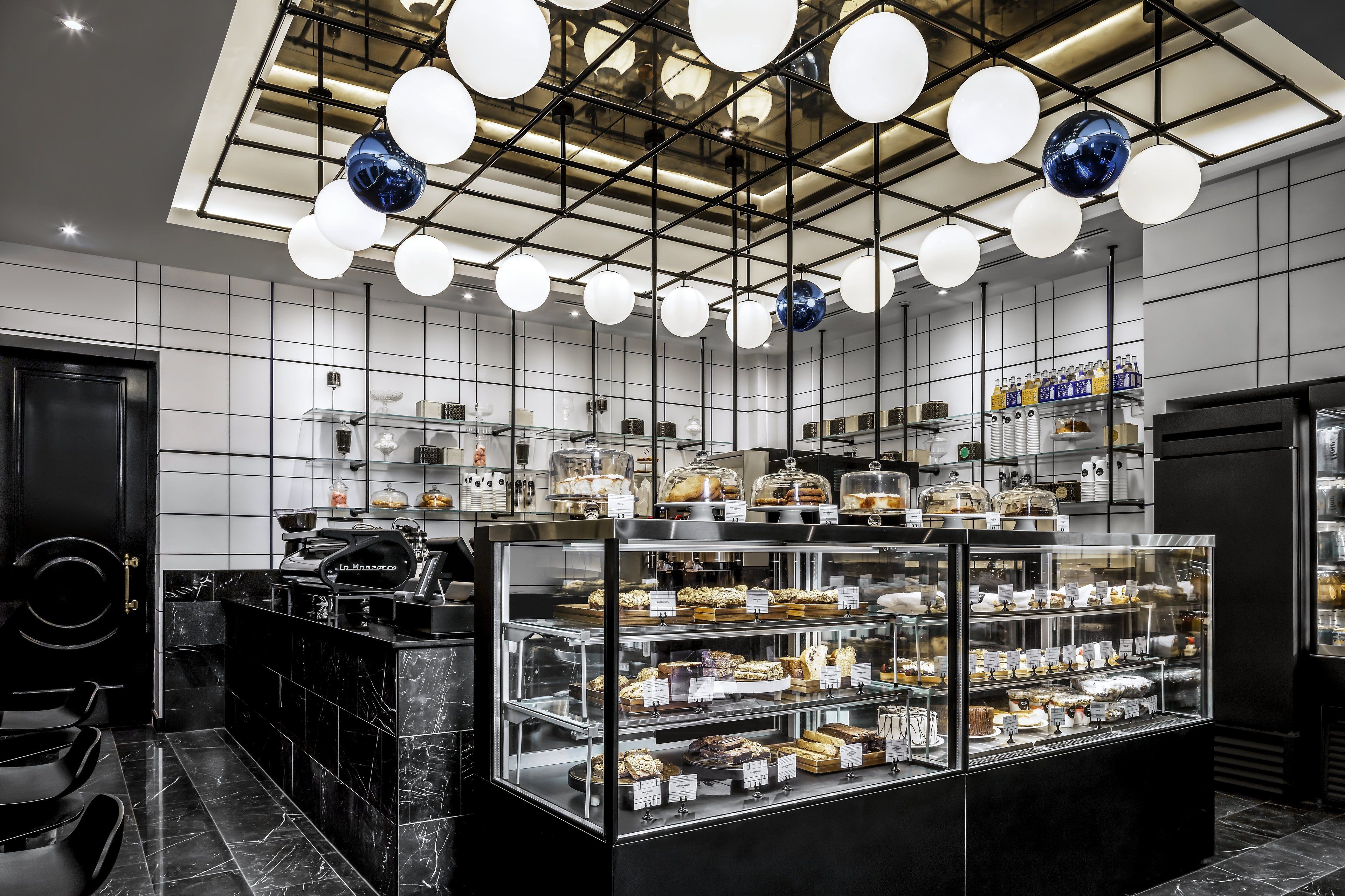 A modern bakery with a glass display case filled with pastries and cakes, a coffee machine, and a seating area with chairs.