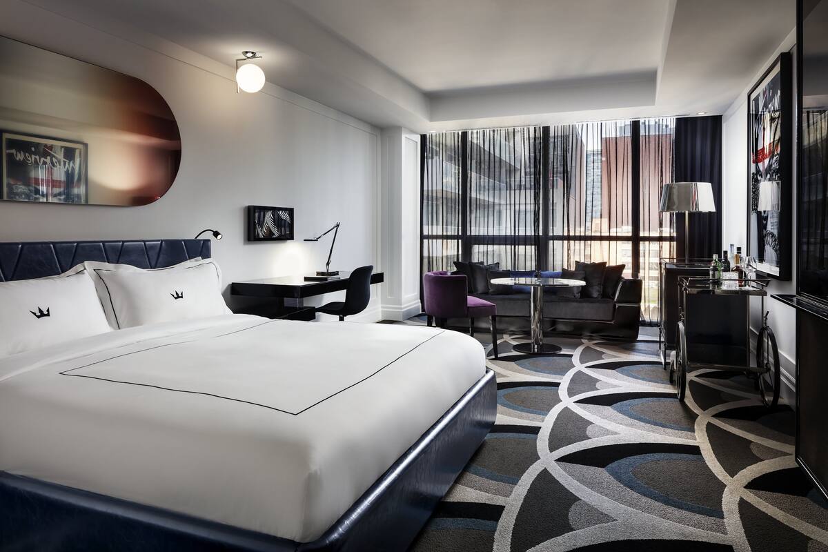 A modern hotel room with a large bed, a desk, a chair, and a view of the city through large windows.