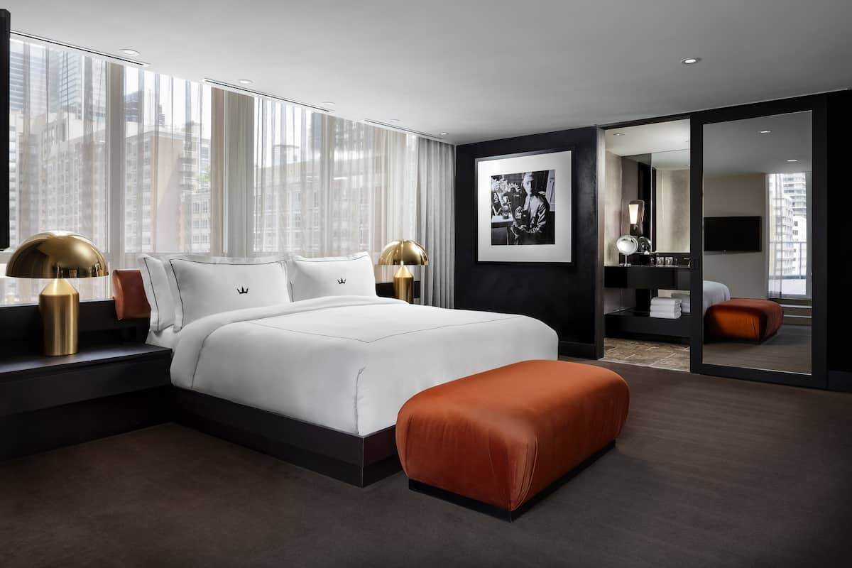 A modern hotel room with a large bed, a sitting area with a red ottoman, and a large window offering a city view.